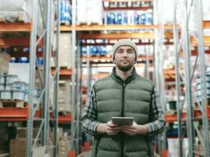 A man holding a tablet stands in a warehouse full of shelves facing the camera.